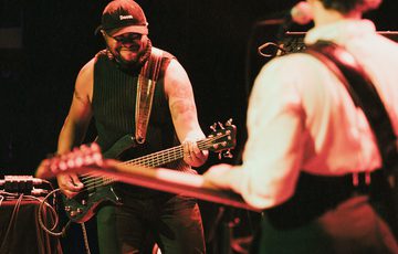 Bassist Neil Charles gives a smiles as he plays his instrument on stage. Neil is wearing a black tank top and a baseball cap with the word 'doom' embroidered on it.