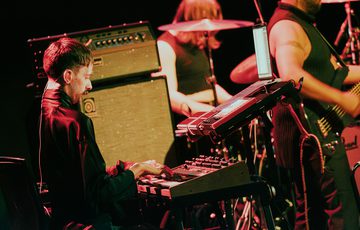 Oliver Vibrans plays keyboard on stage with All Men Unto Me, seated in front of a large amp and surrounded by an array of electronic instruments. Oliver is a young man with short brown hair and a moustache wearing a black shacket.