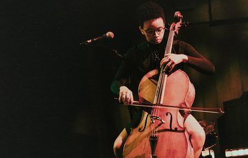Cellist Simone Seales plays their instrument from a seated position, looking down intently. Simone is a young non-binary person wearing glasses and a black polo neck, with short natural curly hair.