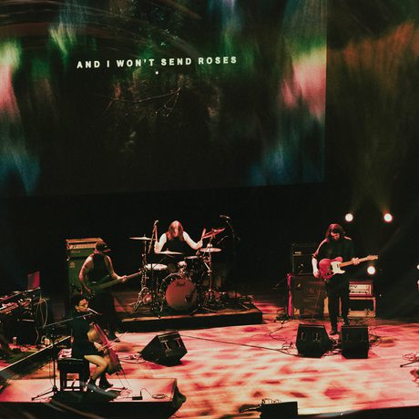 A wide shot of All Men Unto Me performing on stage at Queen Elizabeth Hall, Southbank Centre. They are a six-piece group consisting of drums, bass, cello, electronic, guitars and vocals. Behind them integrated captions are projected showing the lyrics "and I won't send roses"