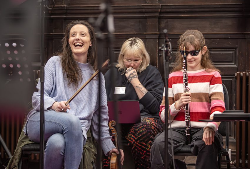 Three musicians are sitting in a rehearsal room. They are laughing and smiling.
