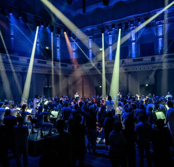 Paraorchestra perform in a large, high-ceiling concert hall illuminated in a deep purple hue with white spotlights criss-crossing the space. Musicians are elevated on small plinths and the audiences are moving amongst them as they play.