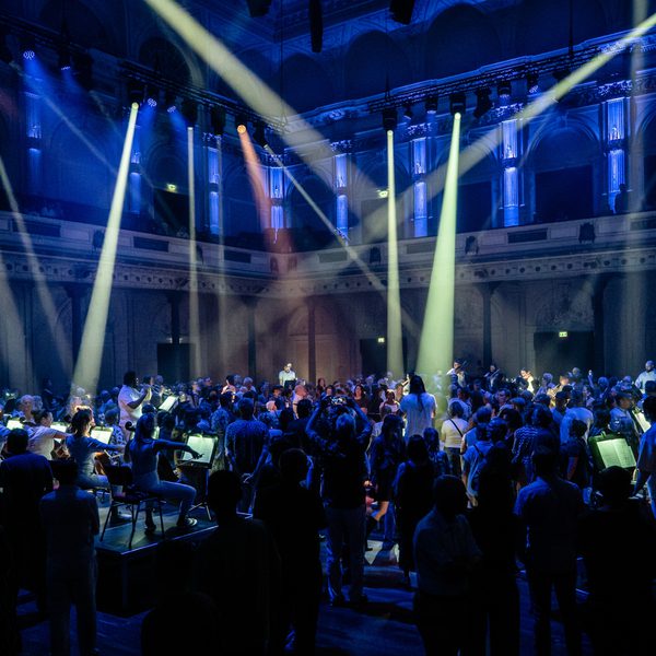 Paraorchestra perform in a large, high-ceiling concert hall illuminated in a deep purple hue with white spotlights criss-crossing the space. Musicians are elevated on small plinths and the audiences are moving amongst them as they play.