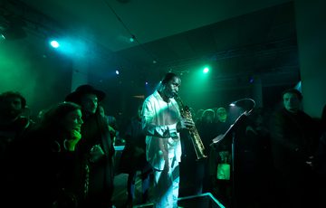 A man with dark skin wearing a white shirt and trousers is playing the tenor saxophone. He is standing on a raised platform while audience members surround him. The room is dark and the performer is illuminated in green hues.