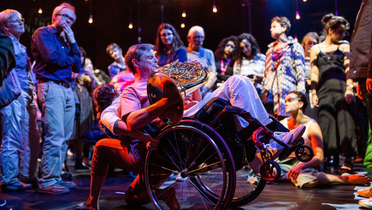 A musician and a dancer are performing with audience members surrounding them in a circle, watching the performance intently. The musician is playing a horn, wearing a pink t-shirt, blue jeans and is a wheelchair user. Their wheelchair is balancing on its back wheels, lifted into the air and being supported by the dancer, who is faced towards the audience. The lighting is atmospheric and the performers are illuminated in contrasting blue and pink hues.