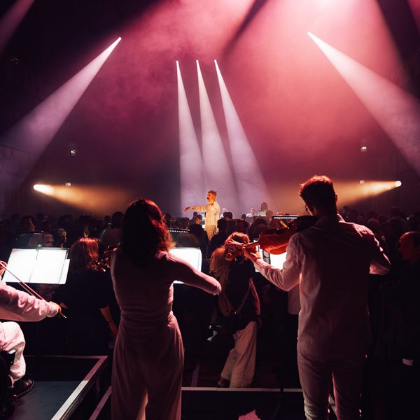 A conductor stands in the centre of a large circle of musicians with his arms outstretched in front of him. He has short hair and a moustache and is wearing all white. The musicians are also all wearing white, and they are playing a variety of instruments including violins and violas. A large audience are interspersed amongst the performers. The room is dark and the performers are illuminated in hues of pink, with beams of white light spotlighting the conductor.