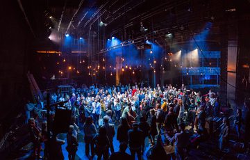 Inside a large theatre, audience and performers mingle together on a stage lit by dozens of filament bulbs suspended from the ceiling. Amongst the crowd a harp, double bass, and other string instruments are visible.