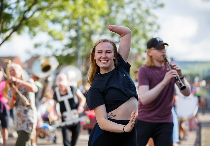 Musicians and dancers rehearse outdoors for SMOOSH! in late 2020. The subject of the photo is a dancer with long red hair and a limb difference, smiling as she strikes a pose.