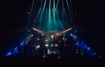 A wide shot of a large concert hall, dramatically lit with green-white spotlights from a high ceiling. Musicians are dotted throughout the space on platforms as the audience walk amongst them.