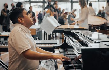 A pianist with short dark hair and medium light skin tone wearing a white polo shirt performs in a bright space with percussionists and audience members visible behind him.