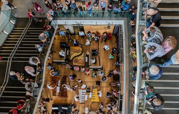 A top-down view showing around a dozen Paraorchestra musicians playing percussion and piano. Audience members lean over to look down from staircases and mezzanines above.