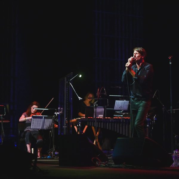 Brett Anderson, a man with light skin a short, dark hair is wearing a black shirt and trousers. He is performing on stage, singing into a microphone. Four musicians are performing with him, including a double bassist, a cellist and a violinist. The stage is dark and the backdrop is black.