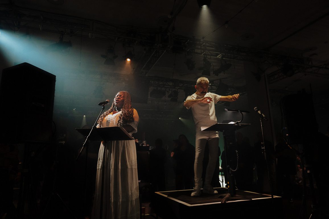 A singer and conductor share a small stage together, illuminated with a single spotlight.