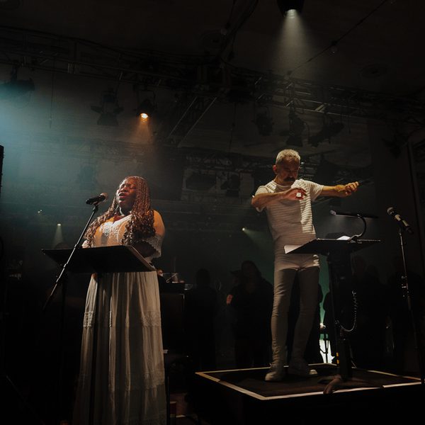 A singer and conductor share a small stage together, illuminated with a single spotlight.