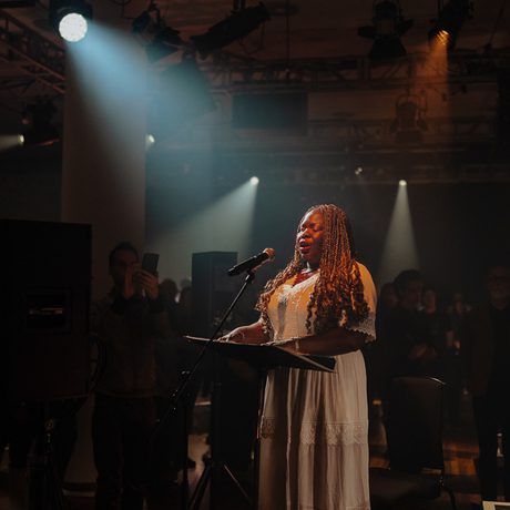 Victoria Oruwari, a woman wearing a white dress with long golden braids and medium ark skin tone, reads braille music as she sings.