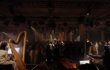 A wide shot of a performance space lit in soft yellow light. Audience members walk amongst musicians positioned on small risers.