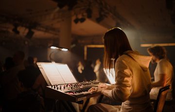 A female keyboard player with light skin tone plays her instrument, her face obscured by her long hair.