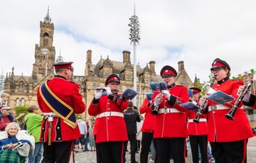Three woodwind musicians from the West Yorkshire Fire Band perform in a public square. They are dressed in traditional red military-style uniforms.