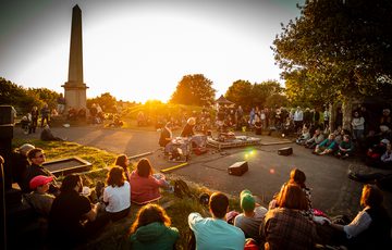 As the sun sets behind them in Undercliffe Cemetery, electronic duo Gilank perform using pedals and midi controllers. Next to them Eeshar Singh plays the santoor, a traditional hammered string instrument. An audience sit on the floor surrounding them.