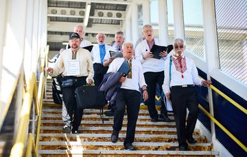 Members of Dibrova Choir, a Ukranian male voice choir wearing traditional embroidered white shirts, sing as they descend a staircase in a train station.