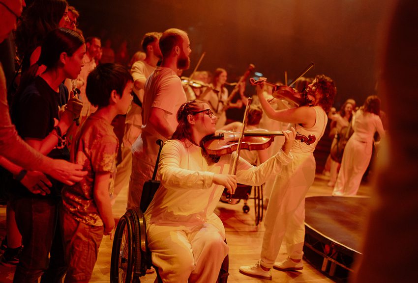 A viola player who is a wheelchair user is at the forefront, surrounded by other musicians and audience members. All of the musicians are wearing white and everyone is dimly lit with a red hue.