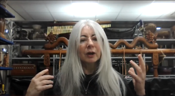 Dame Evelyn Glennie, a person with light skin and long grey hair wearing a black top is talking to a camera on Zoom. They are in a brightly lit space and gesturing with their hands.