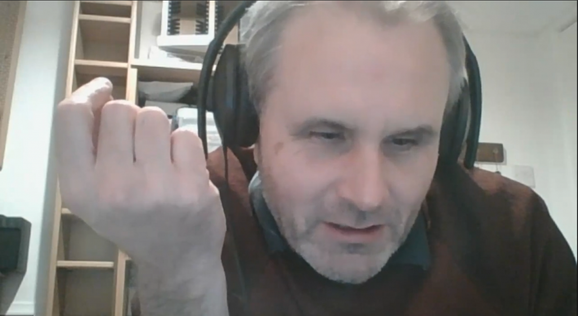 James Risdon, a man with light skin and short grey hair, wearing large black headphones and a brown jumper with a black collared shirt underneath is talking to the camera on Zoom.