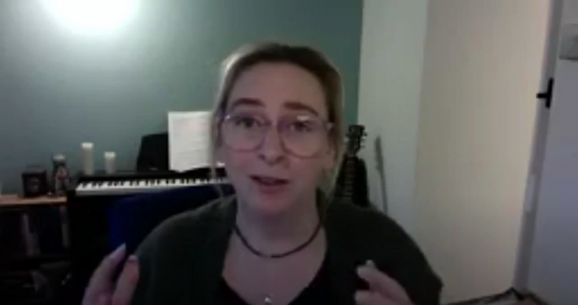 A person with light skin and glasses, wearing a dark cardigan over a black top is talking to the camera on Zoom.