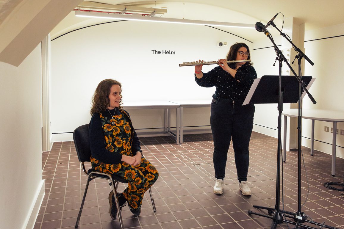 Asteryth Sloane sits on a chair next to Lila Bhattacherjee who is standing up playing a flute. Asteryth is wearing green dungarees with a sunflower motif and a black turtleneck, and has their hands clasped together on their lap. Lila is wearing all black with white trainers, and is reading from sheet music on a stand. There is a microphone in front of them, and they are in a white walled room with a sign that reads The Helm.