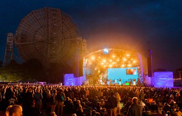 Taken from behind a festival crowd of around 2000 people looking on at a brightly lit stage, nighttime. To the right a huge space telescope. The stage is lit in many yellow spotlights projecting down and out to the crowd. On stage are around 10 musicians. Projections either side of the stage read Bluedot.