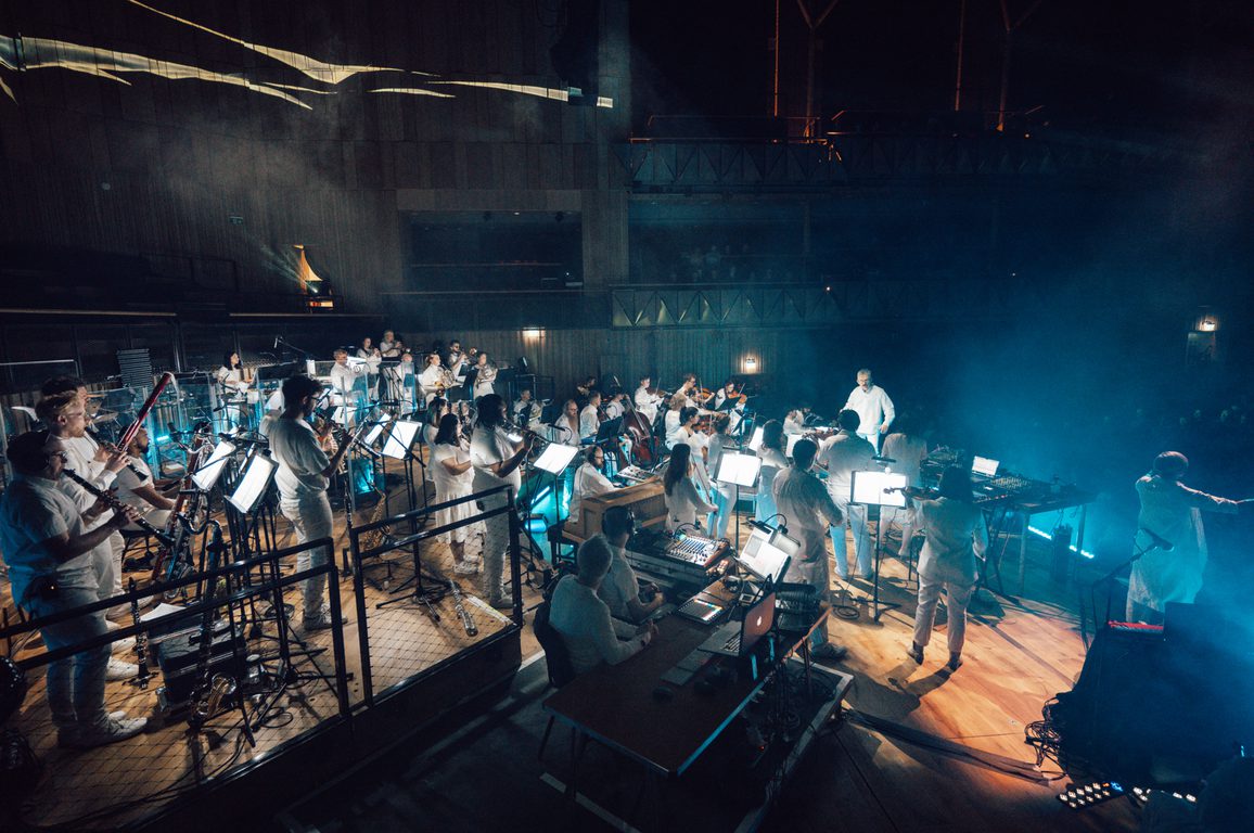 An ensemble of Paraorchestra musicians play on stage with an audience behind them. They are all wearing white and are lit up by bright spotlights.