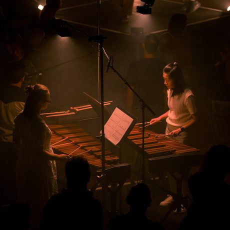 Two marimbists perform facing each other. They are situated on a plinth in a performance space which is dimly-lit in a wash of dark orange lighting.