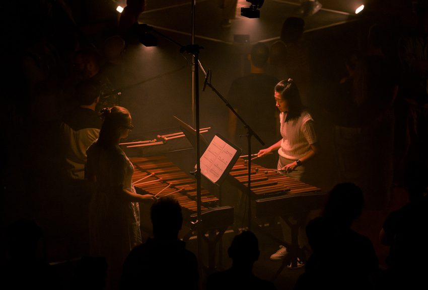 Two marimbists perform facing each other. They are situated on a plinth in a performance space which is dimly-lit in a wash of dark orange lighting.