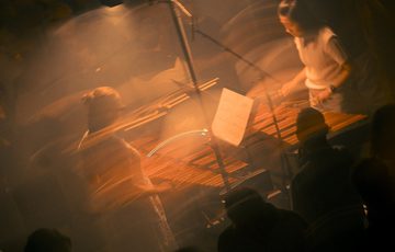 Two marimbists perform facing each other. They are situated on a plinth in a performance space which is dimly-lit in a wash of dark orange lighting. The image has been edited to give a blurred effect, creating a sense of dynamism and motion.