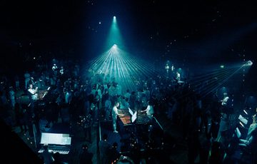 Paraorchestra perform at Beacon Hall in Bristol Beacon. The players are situated on plinths on the floor of the hall, while a large audience roams around them. The space is illuminated in a wash of blue lighting.