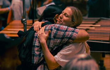 An orchestral player embraces a friend or family member after concluding a performance. In the background of the image, a large audience and a marimba is visible.