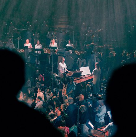 Paraorchesta perform at Bristol Beacon. The musicians are spread across the floor of Beacon Hall, situated on plinths. The hall is lit in a wash of blue lighting, with warm orange spotlights illuminating the players. This photograph is taken from the balcony, with the silhouettes of two audience members in the foreground of the image.