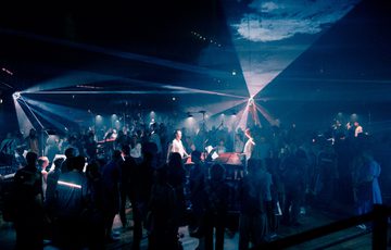 Paraorchestra perform at Beacon Hall in Bristol Beacon. The players are situated on plinths on the floor of the hall, while a large audience roams around them. The space is atmospherically lit in a wash of blue lighting with white spotlights illuminating the players.