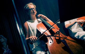 A cellist is performing. She is a woman with blonde hair and glasses, wearing a white top and white trousers. She is sitting down in a performance space which is atmospherically lit in a wash of blue lighting.