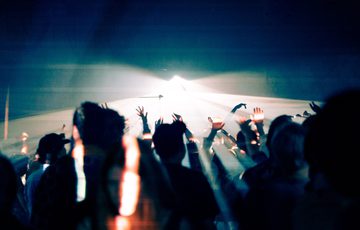 A large audience dances and moves around a performance space. They are illuminated in a wash of blue lighting that contrasts with orange flecks of light. A white spotlight shines through the centre of the audience.