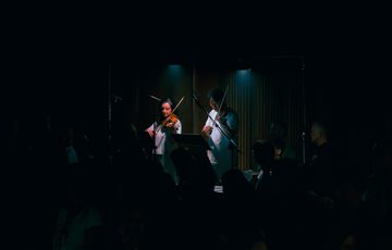 Two violinists, both dressed in white, perform on the floor of Beacon Hall at Bristol Beacon in near darkness. The two players are illuminated by a white spotlight.