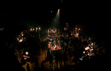 Paraorchesta perform at Bristol Beacon. The musicians are spread across the floor of Beacon Hall, situated on plinths. The space is dimly lit with the players illuminated in warm orange spotlights.