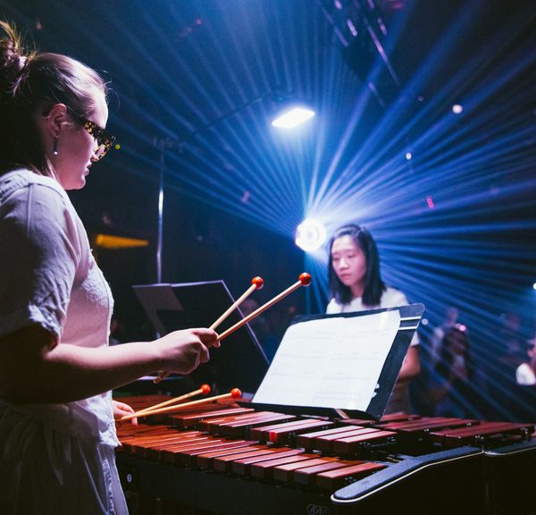 Two percussionists are playing the marimba. They are positioned opposite each other, with two marimbas in between them. They are in a performance space which is illuminated in a radiant blue spotlight with contrasting small specks of orange lights.