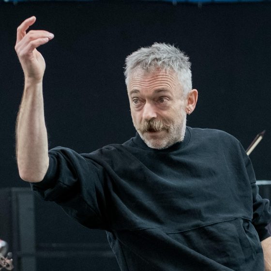 Charles Hazlewood, a man with light skin and short hair wearing a black jumper and light blue jeans, is standing in the centre of a rehearsal space conducting four musicians who are surrounding him. He is focused and his right arm is raised in the air, motioning to the musicians. The rehearsal space is brightly lit.