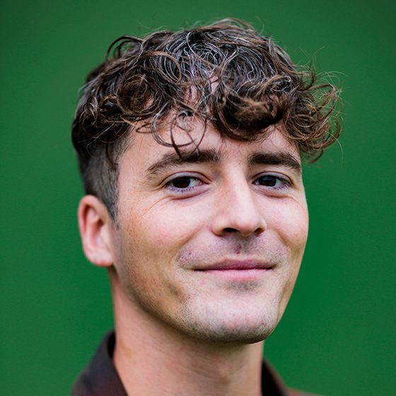 Ben Horton, a man with medium-light skin tone, short brown curly hair and a closed, warm smile. The background behind him is green.