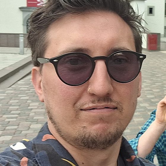 Dean Sudders, a white man in his late 30s is smiling at the camera. Dean has short brown hair which sweeps backwards, a nose piercing and is wearing sunglasses and an abstract patterned shirt.