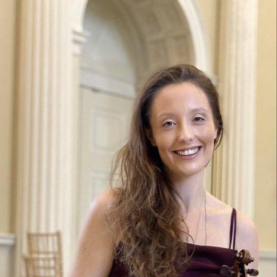 Siobhan Clough, a woman with light skin tone and long brown hair holding her violin and smiling warmly