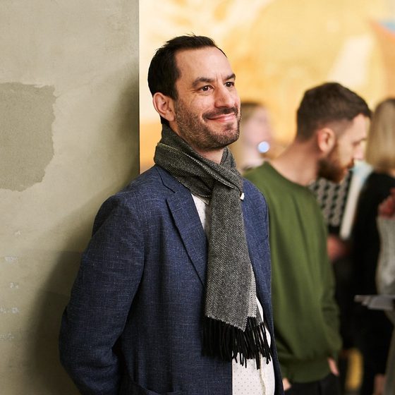 Jonathan Harper, a man with medium-light skin tone, short dark hair and stubble. He wears a navy blue suit jacket and a scarf and is leaning against a pillar, smiling.