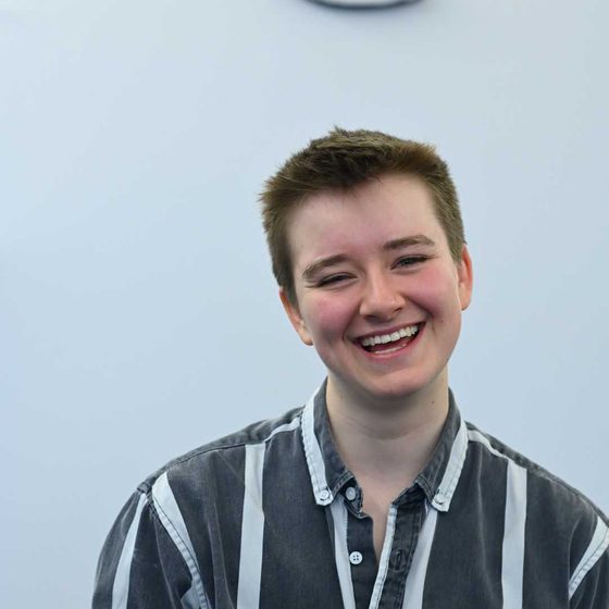 Kit McGrath, a person with short brown hair wearing a striped black and white collared shirt, is smiling animatedly at the camera.