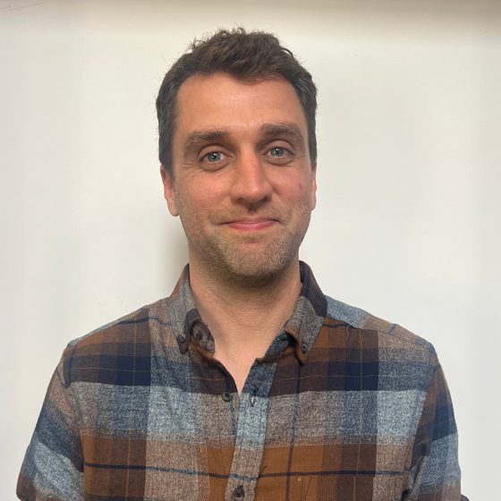 Michael Graham-Court, a man with light skin tone, blue eyes, and short, dark brown hair, is smiling at the camera. He is wearing a light blue, navy blue, and brown plaid flannel shirt.